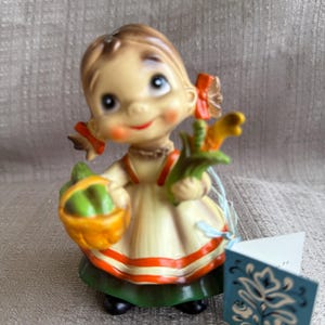 May include: A vintage figurine of a girl with brown hair wearing a white dress with red and orange stripes and a green skirt. She is holding a basket of green vegetables and a bunch of yellow corn. The figurine has a tag with a blue and white floral design.