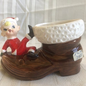 Könnte beinhalten: Ein Keramik-Weihnachtsboot-Pflanztopf mit weißem Pelzbesatz und einer kleinen Elfenfigur, die auf dem Stiefel sitzt. Der Elf trägt einen rot-weißen Weihnachtsmann-Anzug und hat blonde Haare. Der Stiefel hat eine braune Glasur und ein Holzmaserungsmuster.