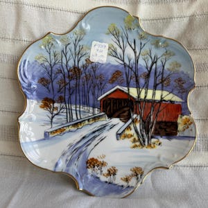 Może przedstawiać: Ozdobny talerz porcelanowy z zimową sceną przedstawiającą czerwony zadaszony most, śnieg i nagie drzewa. Talerz ma falisty brzeg ze złotym wykończeniem i jasnoniebieską obwódką. Talerz ma tekst "P739 4/4 60.00".