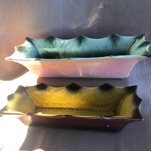 Peut inclure: Deux jardinières rectangulaires en céramique. La jardinière supérieure est rose clair avec un bord vert. La jardinière inférieure est jaune avec une base rouge foncé et un bord noir. Les deux ont un bord festonné.