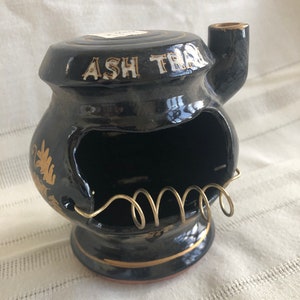 May include: Black ceramic ashtray shaped like a stove with a gold trim and a gold wire grate. The ashtray has a small spout on the side and says "ASH TRAY" in gold lettering.