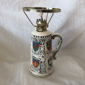 May include: A white ceramic oil lamp with a metal wick holder and a handle. The lamp is decorated with four shields with the words "Loyalty", "Trust", "Faith", and "Hope" written on them.