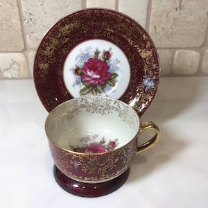 May include: A vintage teacup and saucer set with a burgundy and gold floral design. The cup and saucer feature a delicate floral pattern with a single rose in the center. The saucer has a gold rim and a burgundy background.