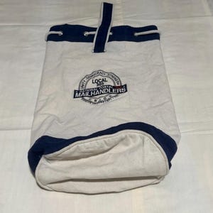 May include: White canvas mail bag with navy blue trim and handle. The bag has a drawstring closure and a circular emblem with the text "LOCAL 305 NATIONAL POSTAL MAILHANDLERS UNION".