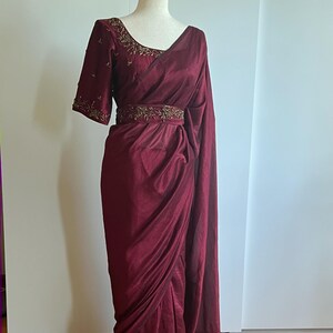 May include: A burgundy saree with a matching blouse and belt. The blouse has short sleeves and gold embroidery around the neckline and sleeve cuffs. The belt is also adorned with gold embroidery. The saree is draped over a mannequin.