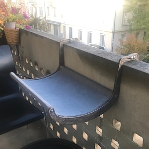 May include: A balcony-mounted shelf featuring a curved, dark-finished metal frame and a smooth, light blue fabric surface. The structure is designed to hook securely over a concrete railing, providing a stable, horizontal platform for various items.
