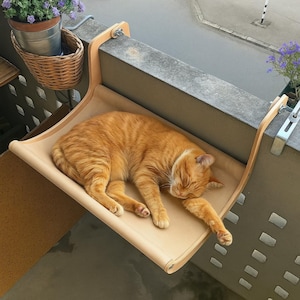 Könnte beinhalten: Eine orangefarbene Katze schläft friedlich auf einer beigefarbenen Katzenhängematte, die an einem Balkongeländer befestigt ist. Die Hängematte besteht aus hellfarbenem Stoff und Holz. Ein geflochtener Korb mit einer Topfpflanze hängt in der Nähe und fügt Grün hinzu.