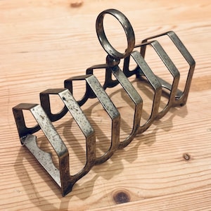 May include: A vintage metal toast rack with six compartments. The rack is made of a tarnished metal, possibly brass, with a ring handle at the top. The rack is angled, with each slot slightly higher than the last.