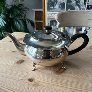 May include: A polished silver teapot with a black handle and lid knob. The teapot has a decorative scalloped edge and rests on three small feet. The teapot is on a wooden surface.