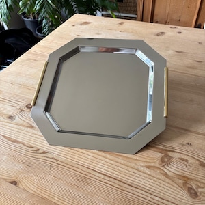May include: A silver-toned octagonal serving tray with a reflective mirrored surface and two vertical gold-toned metal handles attached to the sides. The tray features a smooth, polished finish and a geometric shape with clean, straight edges.