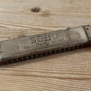 May include: A rectangular harmonica with a weathered silver-toned metal cover plate featuring the embossed text ECHO BELL METAL REEDS. The instrument has a dark brown body with visible air holes along the side and metal rivets at the ends.