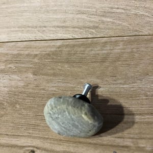 May include: A grey stone cabinet knob with a silver screw and black base. The knob has a natural, textured appearance, and is designed for use on drawers or cabinets. The stone is a light grey colour.