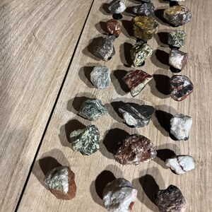 May include: A selection of small rocks and stones in various colours and textures, arranged on a light brown wooden surface. The stones, of varying sizes and shapes, display colours such as white, grey, brown, and touches of red and green. Shadows are visible.