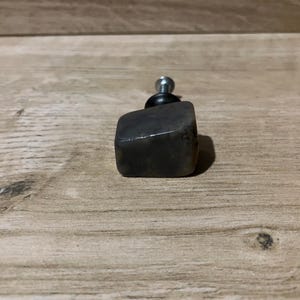 May include: A dark grey, square cabinet knob with a silver metal screw. The knob is made of smooth, polished stone. The knob is set against a light brown wooden surface.