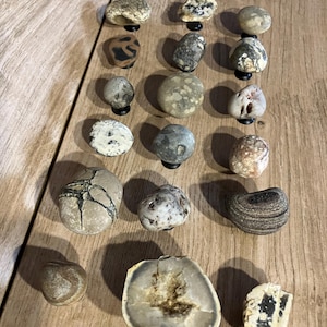 May include: A selection of natural stone cabinet knobs in shades of grey, brown, and beige. The knobs display diverse patterns and textures. Some are round, others are irregularly shaped, offering a rustic aesthetic for furniture.
