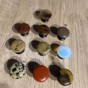 Może przedstawiać: Kolekcja dziesięciu kamiennych gałek do szafek w r&oacute;żnych kolorach i wzorach. Gałki są okrągłe z czarnymi podstawami i zawierają r&oacute;żne kamienie naturalne, takie jak jaspis, tygrysie oko i kamień dalmatyński. Wyeksponowane na drewnianej powierzchni.