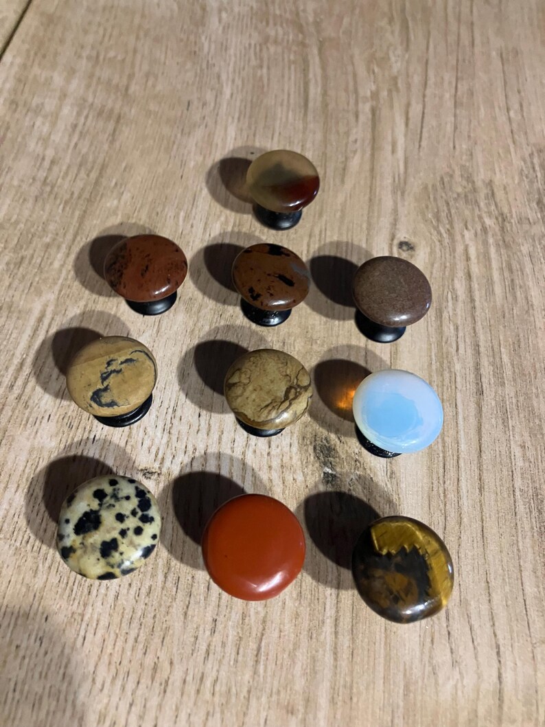 Może przedstawiać: Kolekcja dziesięciu kamiennych gałek do szafek w r&oacute;żnych kolorach i wzorach. Gałki są okrągłe z czarnymi podstawami i zawierają r&oacute;żne kamienie naturalne, takie jak jaspis, tygrysie oko i kamień dalmatyński. Wyeksponowane na drewnianej powierzchni.