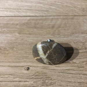 May include: An oval-shaped cabinet knob made from natural stone, featuring a silver-coloured metal screw. The stone has a brown base with a white, cross-like pattern. Set against a wooden background.