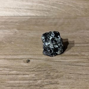 May include: A small, rough-textured rock with a speckled pattern of black and white. The stone is set against a wooden background, showcasing its natural colours and details. This item is a decorative stone.