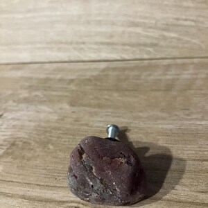 May include: A cabinet knob crafted from a natural, reddish-brown stone. The stone features a textured surface with grey and white veining. A silver-coloured metal screw is attached for mounting. This decorative knob adds a rustic touch to furniture.