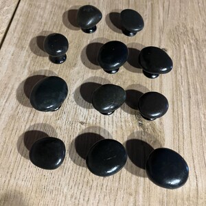 May include: A selection of black, pebble-shaped cabinet knobs with a glossy finish. The knobs are arranged on a light brown wooden surface. Each knob has a small, dark base for mounting.