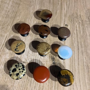 May include: A selection of ten decorative cabinet knobs crafted from polished stones. The knobs display diverse colours and patterns, including red, brown, beige, and speckled designs. Each knob is mounted on a black base.