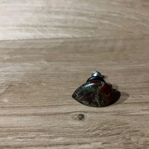 May include: A teardrop-shaped bottle stopper with a polished stone top. The stone has deep red and green hues, with a silver metal top and a black rubber seal. The stopper is designed for a bottle.
