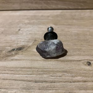 May include: A cabinet knob made from a natural amethyst stone, featuring a silver screw and black base. The knob is a mix of purple and white colours. The knob is on a wooden surface.