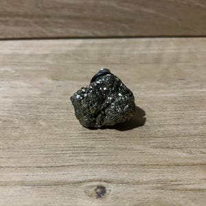 May include: A pyrite gemstone, also known as fool's gold, with a metallic, reflective surface. The pyrite is a dark, shimmering colour and is mounted on a silver-coloured post. The gemstone is displayed on a wooden surface.
