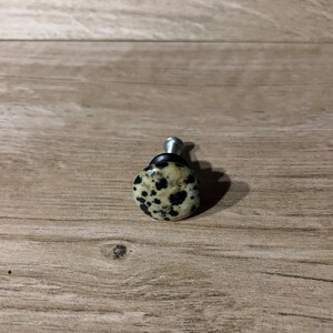 Może przedstawiać: Gałka do szafki z nakrapianą, kremową kamienną powierzchnią i srebrnym metalowym trzpieniem. Kamień ma czarne plamy. Gałka jest przeznaczona do mebli i wystroju domu.