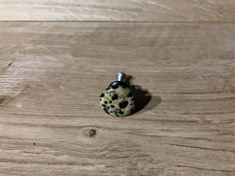 Może przedstawiać: Gałka do szafki z nakrapianą, kremową kamienną powierzchnią i srebrnym metalowym trzpieniem. Kamień ma czarne plamy. Gałka jest przeznaczona do mebli i wystroju domu.