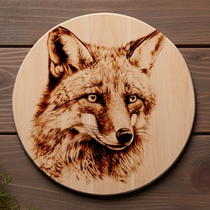 May include: A wood-burned image of a fox's head on a round wooden plaque. The fox has a reddish-brown coat and is looking directly at the viewer.