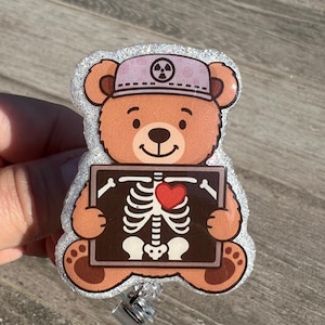 Pode incluir: Um crachá de urso de pelúcia castanho com um chapéu roxo claro com um símbolo de radiação. O urso segura uma imagem de raio-x castanha com um coração vermelho e um esqueleto. O crachá tem uma borda de glitter prateado.