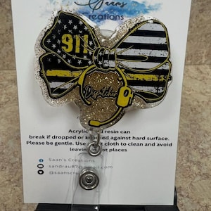 Könnte beinhalten: Ein Ausweis-Rollhalter mit einem Schleifen-Design mit einem gelb-schwarzen amerikanischen Flaggen-Thema, der Nummer 911 und dem Wort "Dispatcher". Die Rolle besteht aus Acryl und Harz mit Glitzerakzenten.