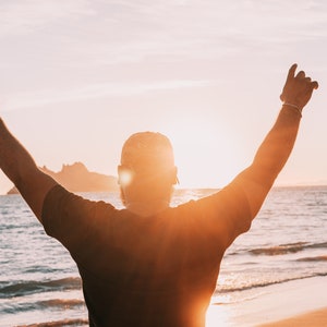 May include: A person with their arms raised in the air, standing on a beach with the sun setting in the background.