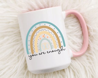May include: White ceramic mug with a pink rim and handle. The mug features a rainbow design with a light blue, yellow, and brown color scheme. The rainbow has a dotted pattern and small flower and butterfly accents. The text "you are enough" is written in black below the rainbow.