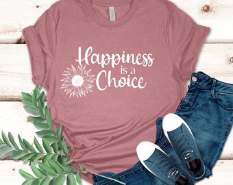 May include: A dusty rose t-shirt with the phrase "Happiness is a Choice" and a sunflower graphic in white. The shirt is paired with blue jeans and navy sneakers with white soles. Green leafy sprigs are in the foreground.