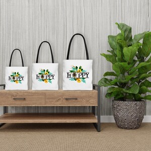 May include: Three white tote bags with black handles and a floral design with bees and the word "Happy" printed on them. The bags are arranged in a row on a wooden shelf.