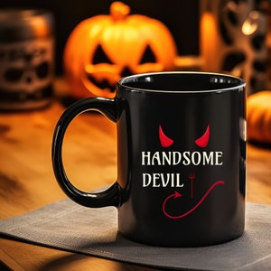 以下が含まれることがあります： 「HANDSOME DEVIL」の白い文字と赤い悪魔の角、赤い三叉と尻尾のデザインが特徴の黒いセラミックマグカップ。 マグカップは湾曲したハンドルを持ち、彫刻されたカボチャのあるぼやけた背景に置かれています。