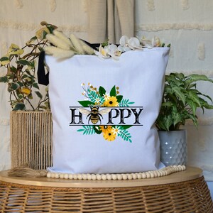 May include: White canvas tote bag with black handles featuring a bee and floral design. The text "HAPPY" is printed in black with a vintage style font.