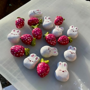 May include: A collection of handmade ceramic ornaments featuring strawberries and bunnies. The ornaments are painted in shades of pink, white, and green.