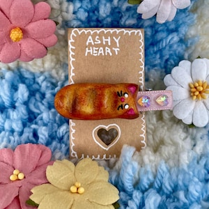 Può includere: Un fermaglio per capelli a forma di gatto arancione fatto a mano con un nastro rosa e dettagli scintillanti. Il fermaglio è su una carta marrone con il testo "ASHY HEART" scritto su di essa.