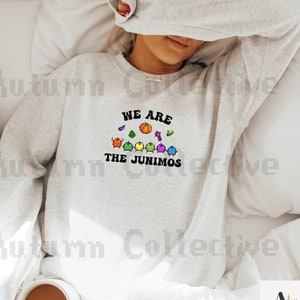 以下が含まれることがあります： 「We are the Junimos」という文字と、ビデオゲーム『Stardew Valley』のカラフルなジュニモのグラフィックが描かれたグレーのトレーナーです。