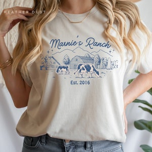 Puede incluir: Una camiseta beige claro con un diseño gráfico azul de una escena de granja. El gráfico incluye una casa de campo, un granero, dos vacas y el texto "Marnie's Ranch Est. 2016".