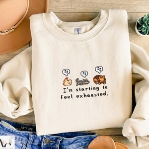 May include: A cream-colored sweatshirt with a pixelated graphic of a chicken, cat, and dog sleeping with the text "I'm starting to feel exhausted."