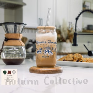 Könnte beinhalten: Ein Glasgefäß mit braunem Deckel und einem blauen und weißen Design mit einer Farm-Szene mit Kühen und dem Text "Nannie's Ranch Est. 2016". Das Gefäß hat einen Strohhalm und steht auf einem Untersetzer aus Holz.