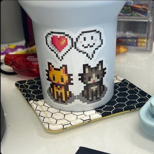 May include: A white ceramic mug with a black and white honeycomb coaster. The mug has four stickers on it: two pixelated cats, one with a red heart and one with a white cloud.