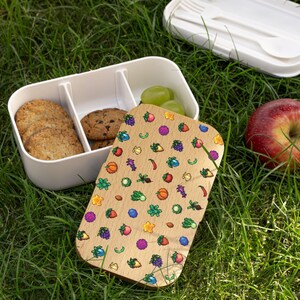 May include: A white lunchbox with a wooden lid featuring a pixelated pattern of various fruits and vegetables. The lunchbox is open and contains cookies and grapes.