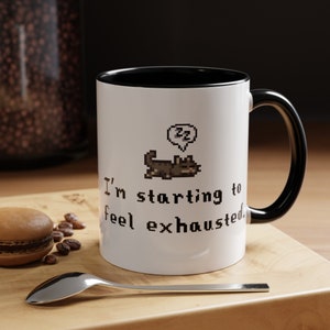 May include: White ceramic mug with a black interior and handle. The mug features a pixelated cat graphic with a speech bubble and the text "I'm starting to feel exhausted.". A spoon, a macaron, and coffee beans are on a wooden surface.