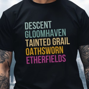 May include: Black t-shirt with a vertical stack of text in a retro color scheme. The text reads "DESCENT", "GLOOMHAVEN", "TAINTED GRAIL", "OATHSWORN", and "ETHERFIELDS".